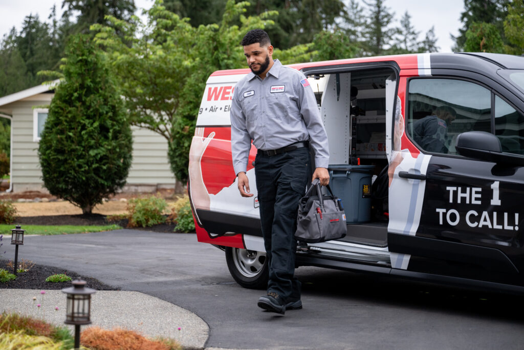 HVAC tech arriving to the customer's home, carrying tool bag from open service truck behind him, parked in driveway.