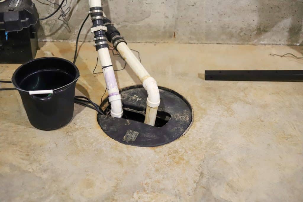 A sump pump system with white discharge pipes is installed in a basement floor pit, with a black bucket and exposed concrete flooring nearby.