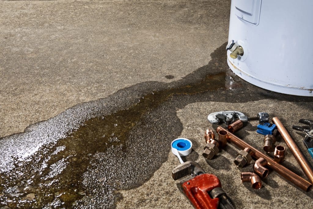 Water leaking from the base of a water heater, forming a large puddle on the concrete floor beside plumbing tools and copper pipe fittings.